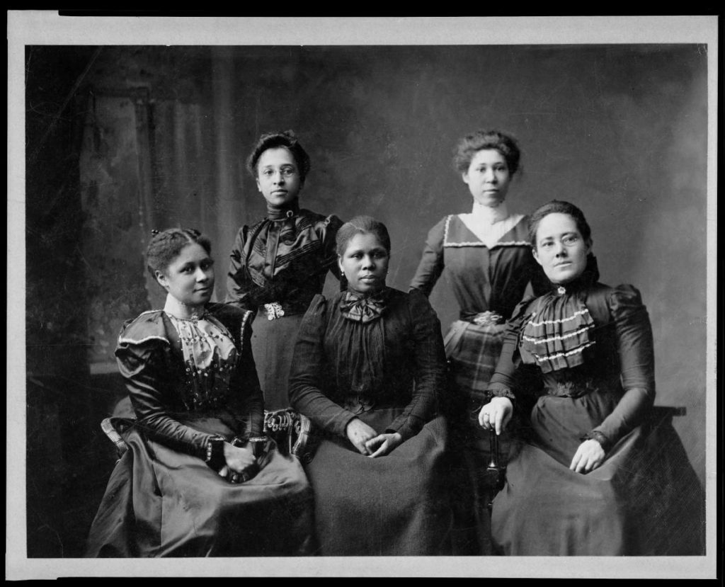 Posed photo of five Black women in formal 19th century dresses.