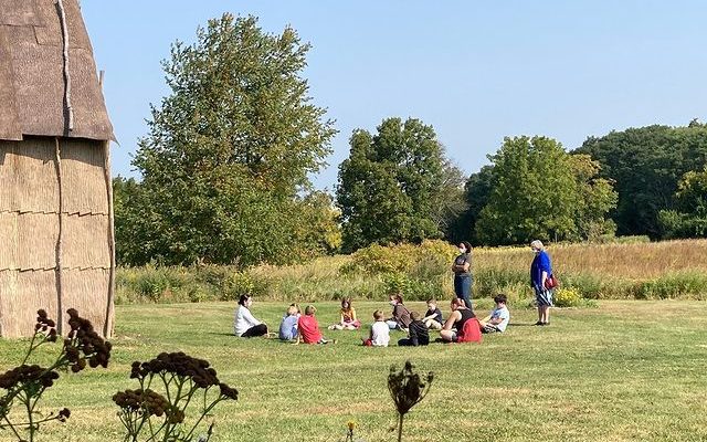 People sit together in a field next to a long house.